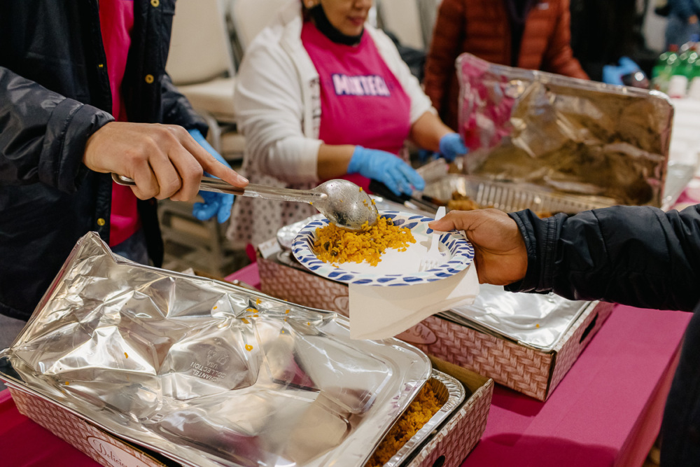 A person serves rice onto a paper plate at a food station, with chafing dishes and volunteers wearing gloves in the background.