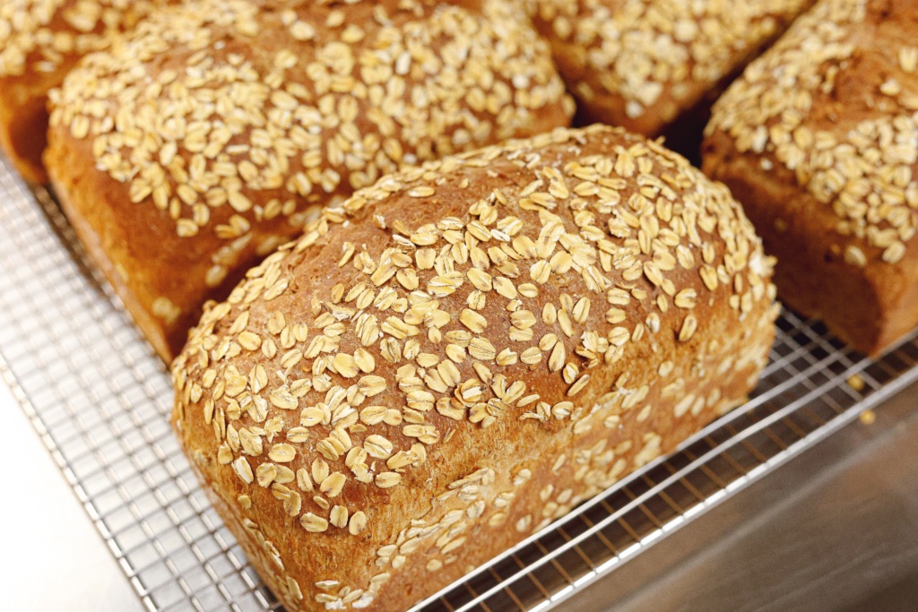 A loaf of oat-topped whole grain bread sits on a cooling rack, with other similar loaves in the background.