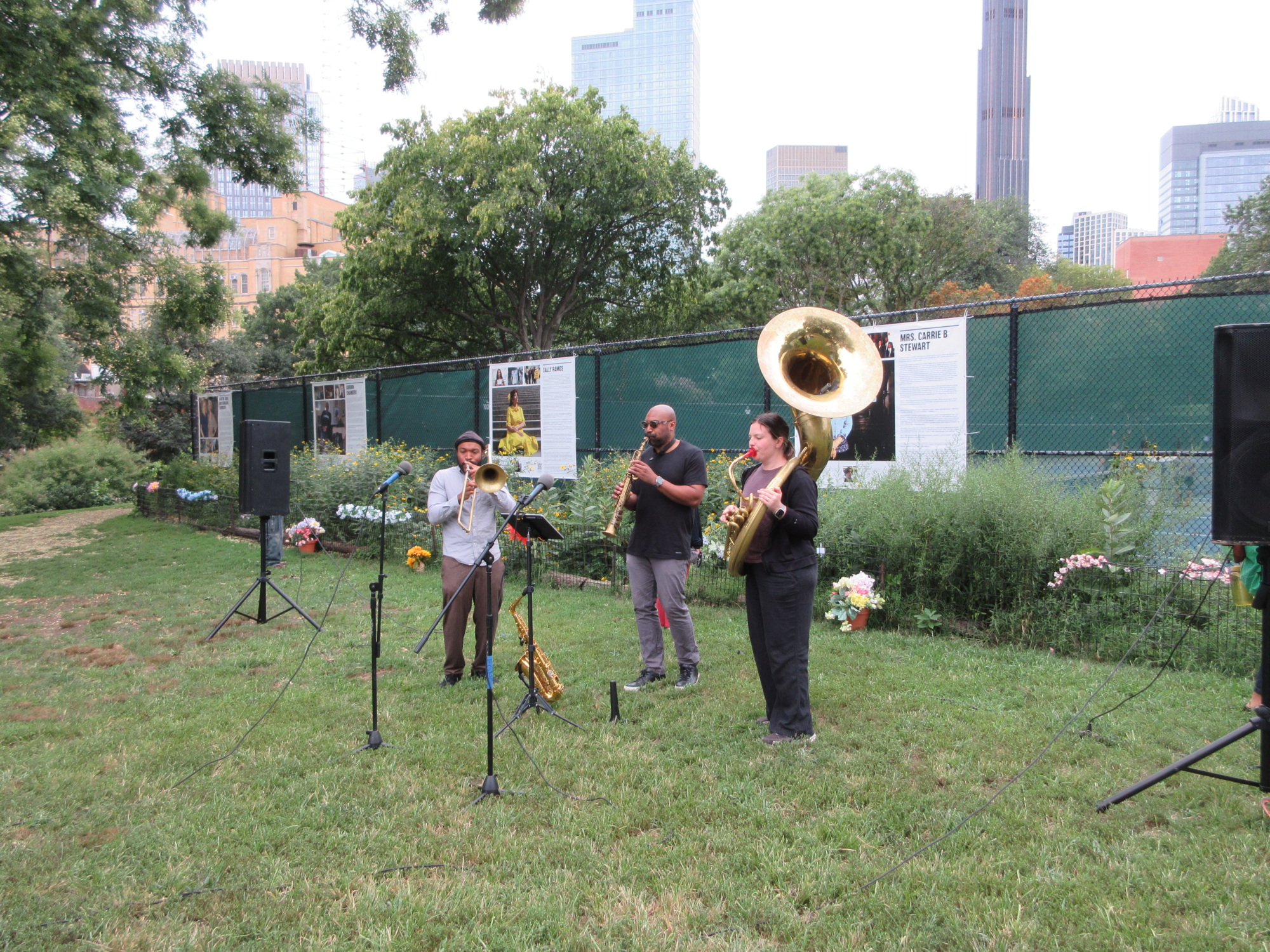 Three musicians play brass instruments outdoors on grass, with city buildings and informational posters visible in the background.