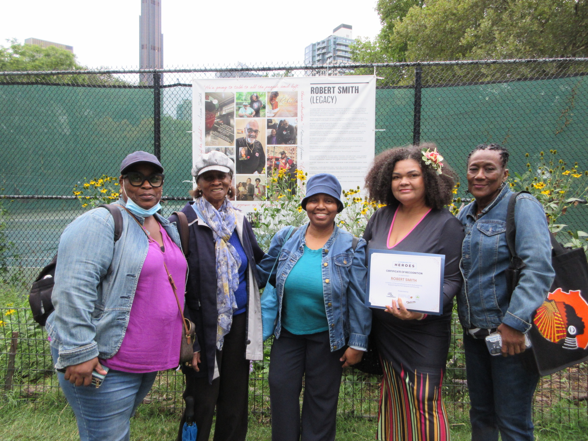 Five women stand in front of a fence with a poster honoring Robert Smith’s legacy, posing for a group photo outdoors. One woman holds a certificate.