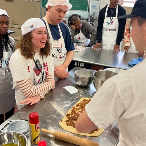 A group of people wearing aprons and hairnets stand around a metal table while one person prepares dough with filling in a kitchen setting.