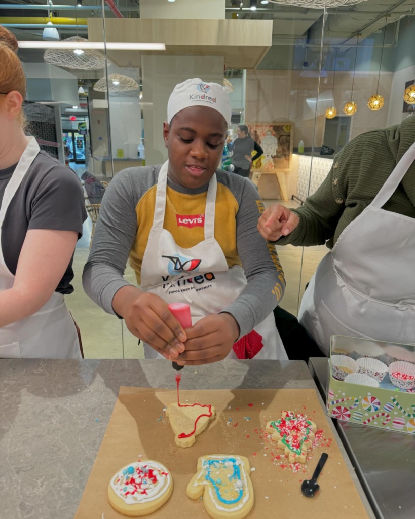 A person wearing an apron decorates cookies with icing at a kitchen counter, surrounded by sprinkles and partially decorated cookies. Two other people are partially visible nearby.