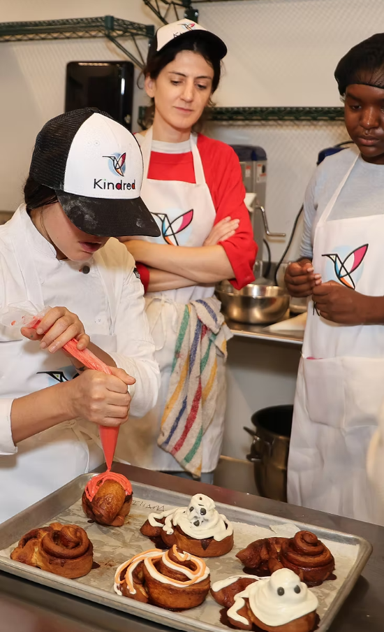 Three people in aprons watch as one decorates cinnamon rolls with icing in a kitchen, with finished rolls on a baking tray.