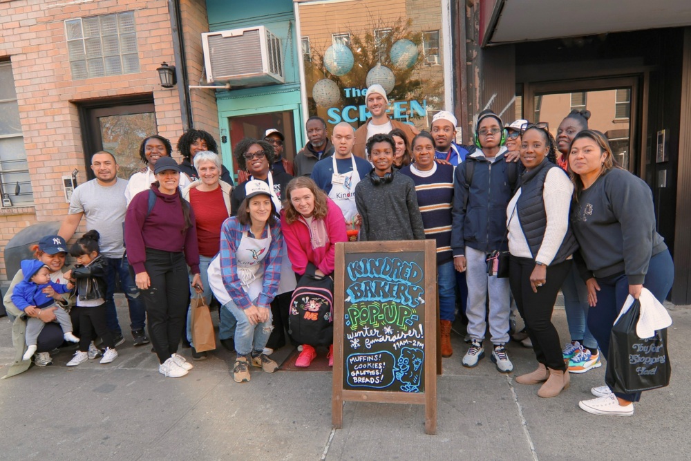 A diverse group of people poses together on a sidewalk in front of a storefront with a chalkboard sign reading "Kindred Bakers Pop-Up.