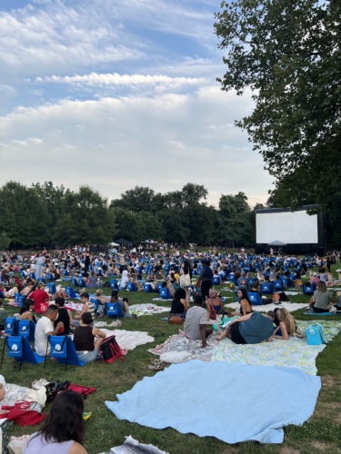 A large crowd sits on blankets and lawn chairs in a park facing a blank outdoor movie screen, surrounded by trees under a partly cloudy sky.