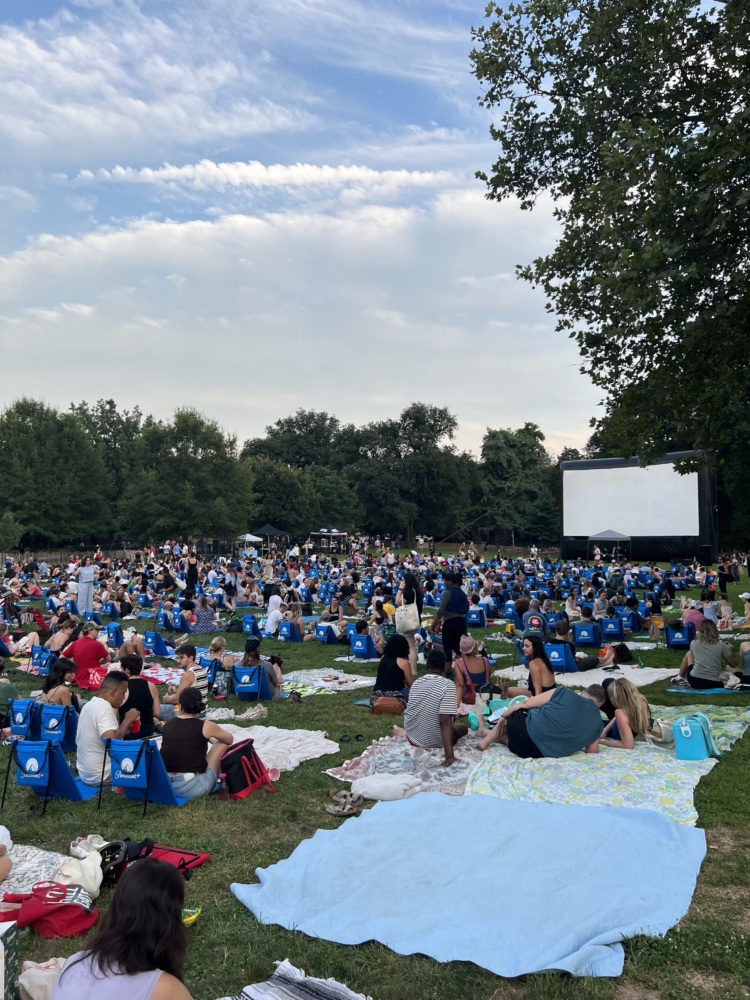 A large crowd sits on blankets and lawn chairs in a park facing a blank outdoor movie screen, surrounded by trees under a partly cloudy sky.
