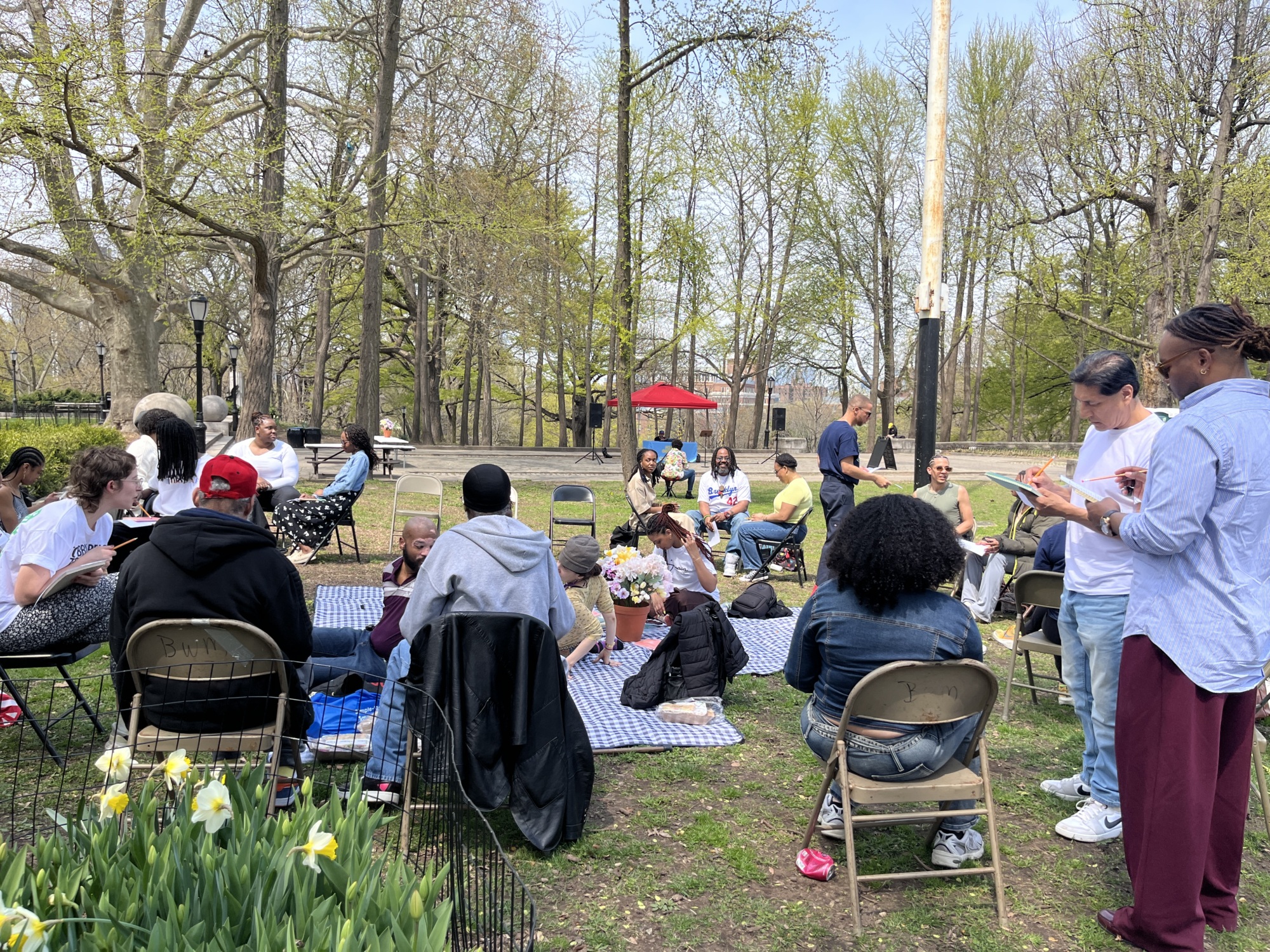 A group of people sit on chairs and blankets in a park, gathered for an outdoor event on a sunny day with trees and a red umbrella in the background.