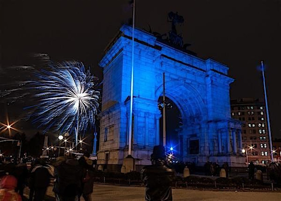 A large stone arch monument is illuminated in blue light at night, with fireworks bursting in the sky and people gathered in the foreground.
