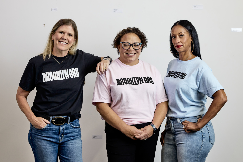Three women stand side by side, smiling, wearing t-shirts that read "BROOKLYN.ORG" in black or white text against a plain background.