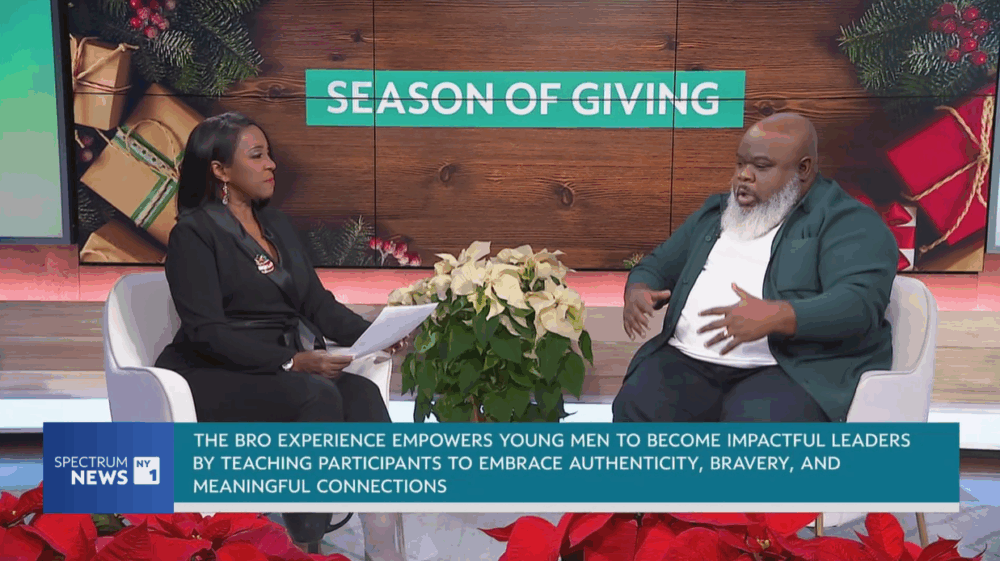 Two people have a discussion on a talk show set decorated with poinsettias and a "Season of Giving" sign, with a caption about empowering young men through leadership programs.