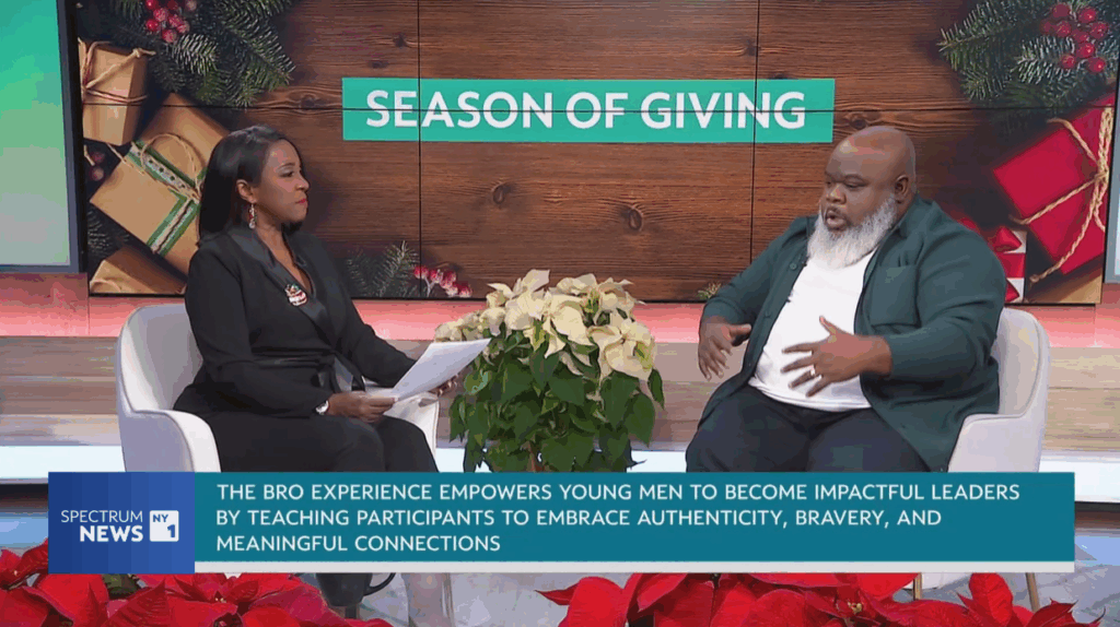 Two people have a discussion on a talk show set decorated with poinsettias and a "Season of Giving" sign, with a caption about empowering young men through leadership programs.