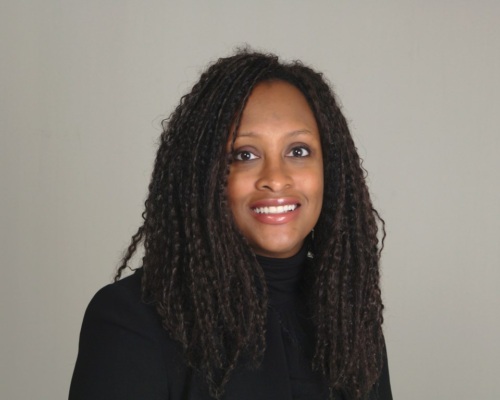 A woman with long curly hair, wearing a black top, smiles at the camera against a plain light-colored background.