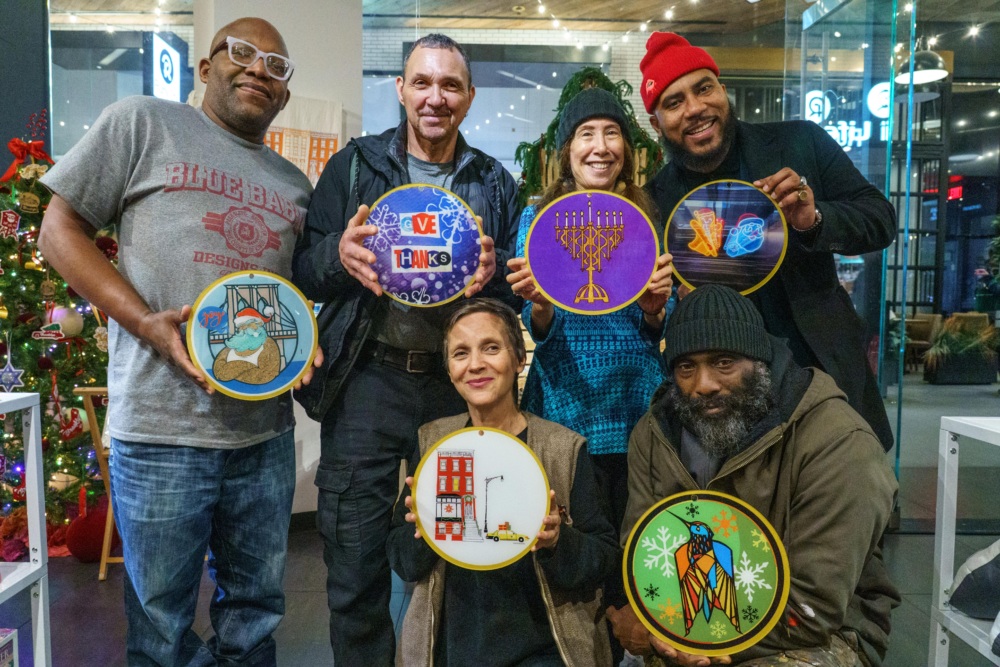 Six people stand indoors, each holding an embroidery hoop with different designs, including holiday, Hanukkah, and winter-themed images. A decorated Christmas tree is visible in the background.