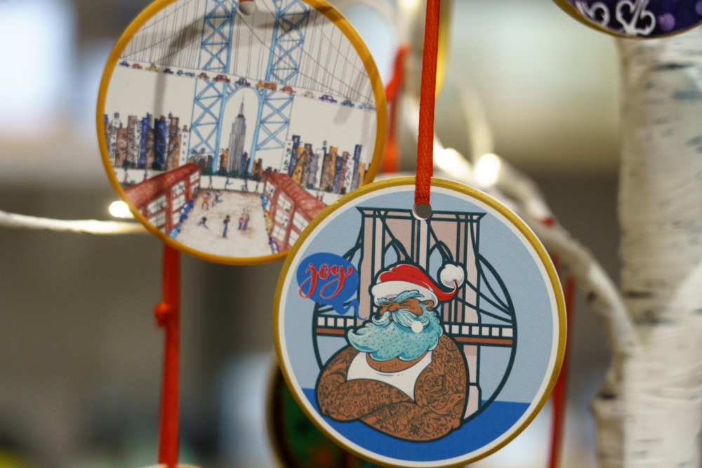 Two round ornaments hang on a tree; one shows a city bridge scene, the other depicts a tattooed Santa Claus in front of a bridge with the word "Joy.