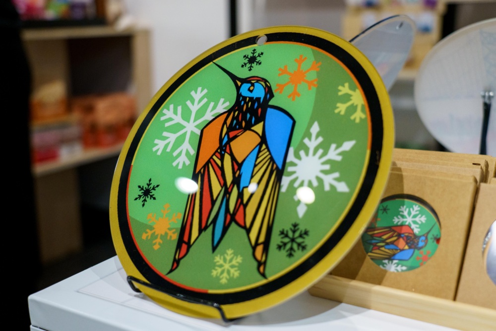 A round decorative plate with a colorful, geometric bird design and white snowflakes on a green background is displayed upright on a stand.