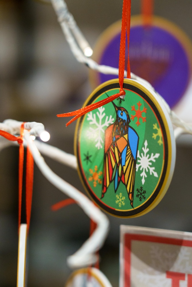 A round ornament with a colorful bird design and snowflakes hangs from a white branch by a red ribbon.