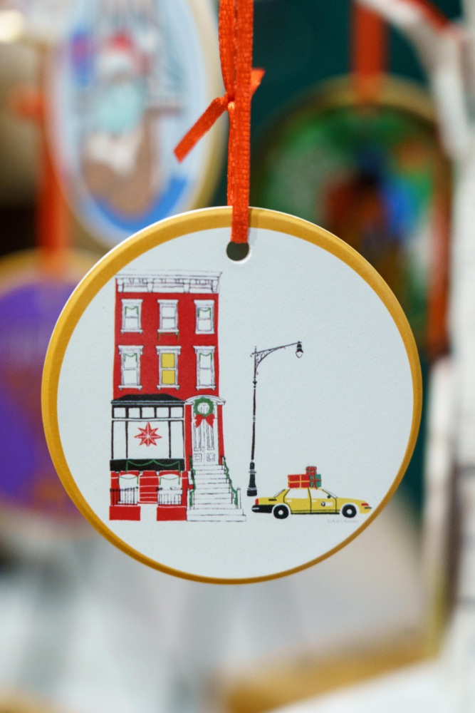 A round ornament decorated with an illustration of a red brick building, a streetlamp, and a yellow taxi with gifts on top, hanging by a red ribbon.