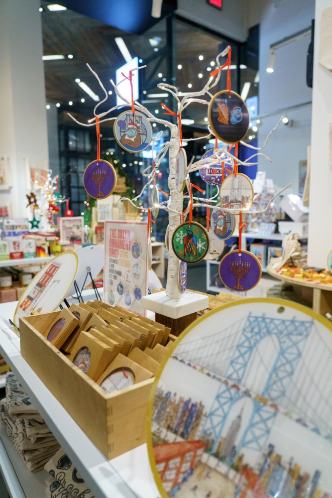 Display of colorful holiday ornaments hanging on a white tree stand, surrounded by various souvenir items and gifts in a well-lit shop.