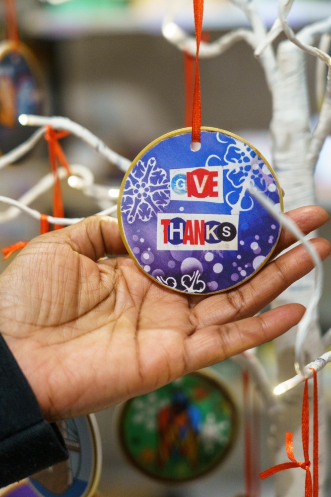 A hand holds a circular ornament with a blue snowflake design and the words "GIVE THANKS" hanging from a red ribbon on a white decorative tree.