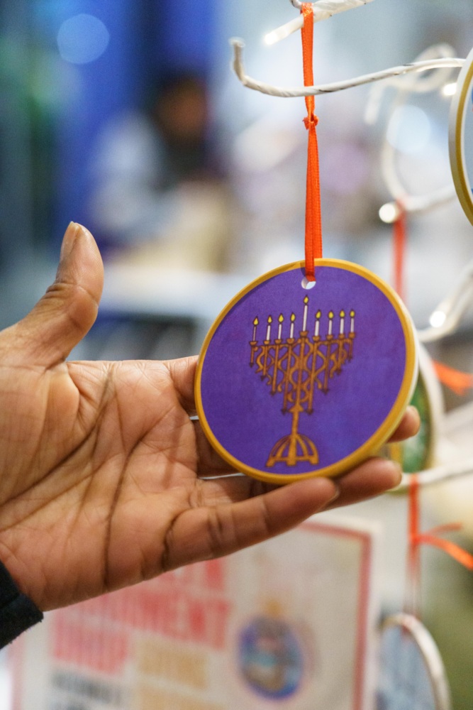 A hand holds a round ornament with a gold menorah design on a purple background, hanging by a red ribbon from a display stand.