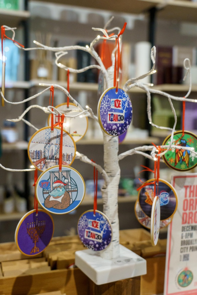 A small white decorative tree displays colorful, round ornaments with various designs and text, set indoors on a shelf.