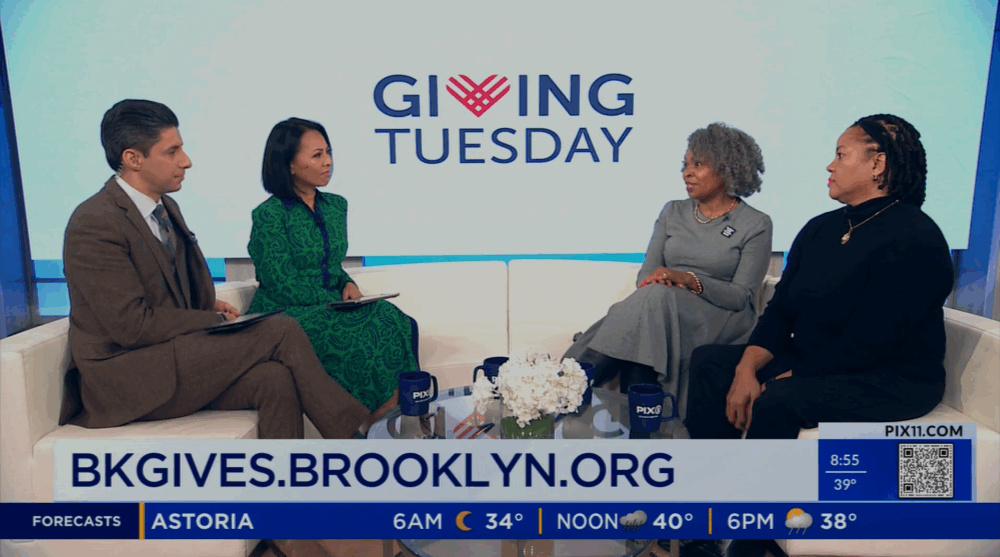 Four people sit and talk on a news set with a "Giving Tuesday" sign behind them and a web address, BKGIVES.BROOKLYN.ORG, displayed at the bottom of the screen.