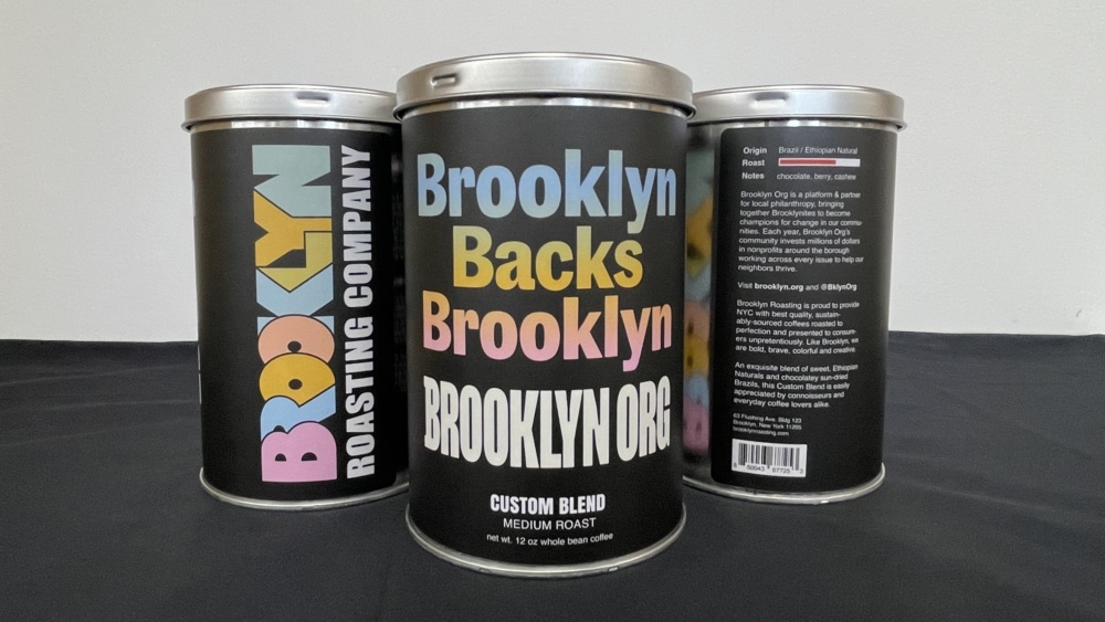 Three cylindrical cans of Brooklyn Roasting Company custom blend coffee are arranged on a black surface, with labels displaying branding and product details.