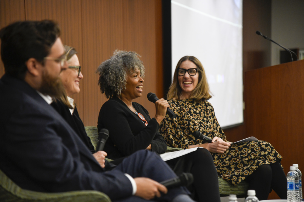 Four people sit on a panel in front of a wooden wall, with one woman speaking into a microphone while the others listen and smile.