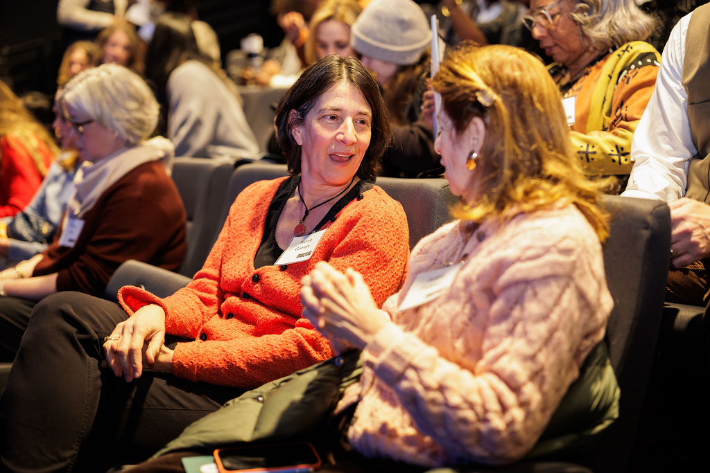 Two women sit in an auditorium, engaged in conversation, while other people are seated around them, some talking and some listening.