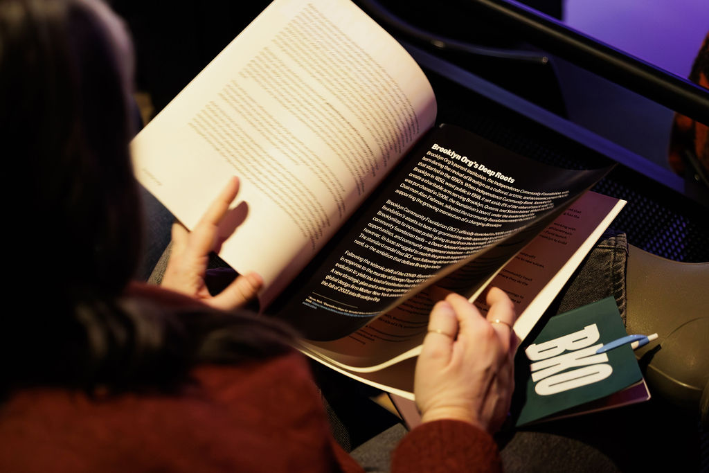 A person reads a booklet or program, with a visible page titled "Bringing Off-Page Reads." A green badge labeled "BIO" and a pen rest nearby.