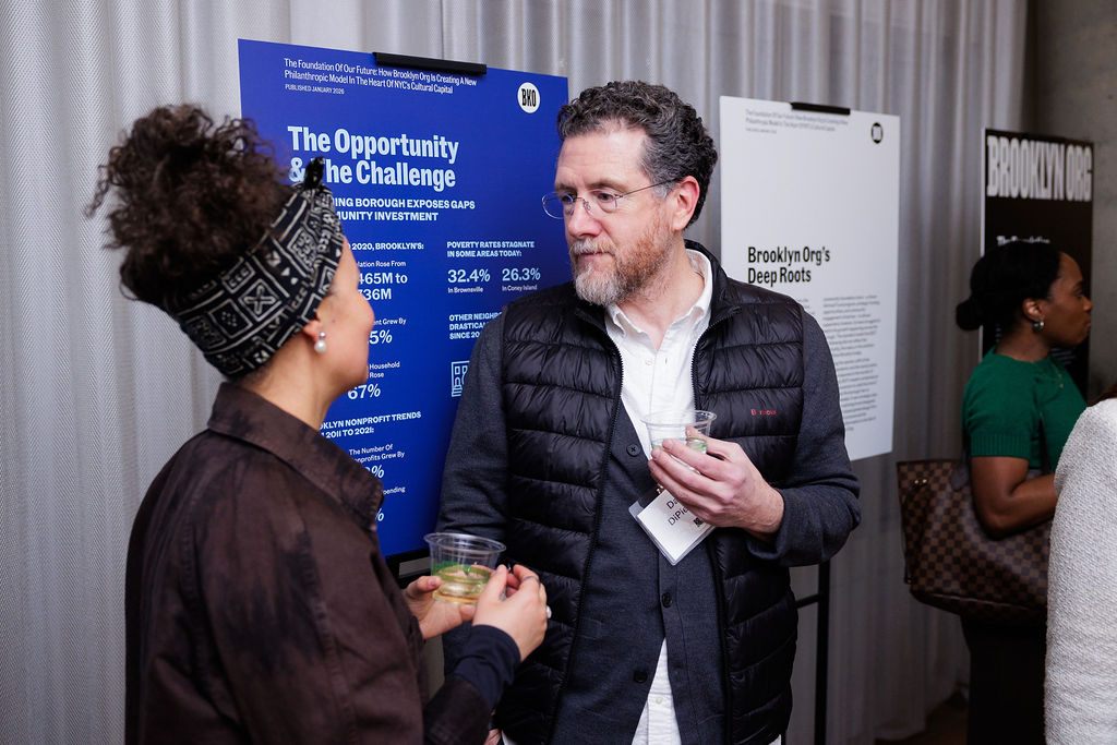 A man and woman converse while holding drinks in front of informational posters at an indoor event.