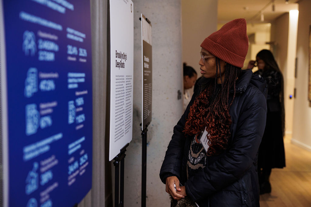A person in a red beanie and winter coat stands indoors, reading information displayed on a vertical board at an exhibition.
