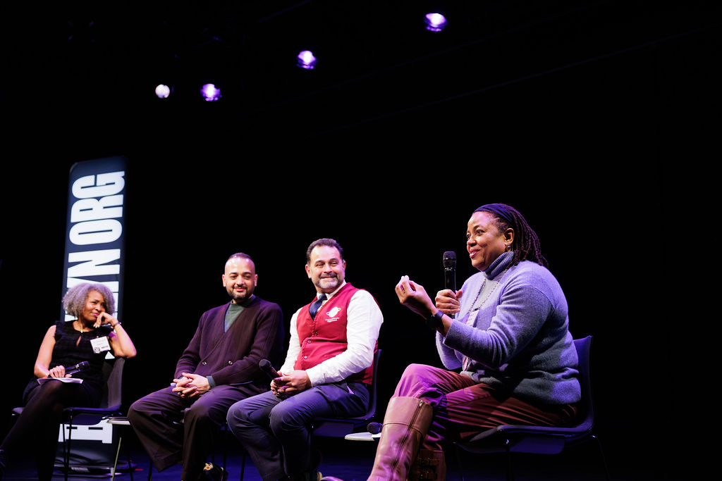 Four people sit on stage under spotlights, three listening as one woman speaks into a microphone. A large vertical sign reading “BROOKLYN ORG” is visible in the background.