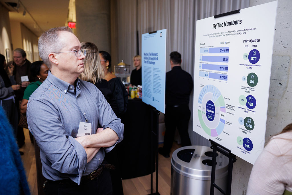 A man with glasses stands with arms crossed, looking at an infographic display titled "By The Numbers" at an indoor event with other attendees in the background.