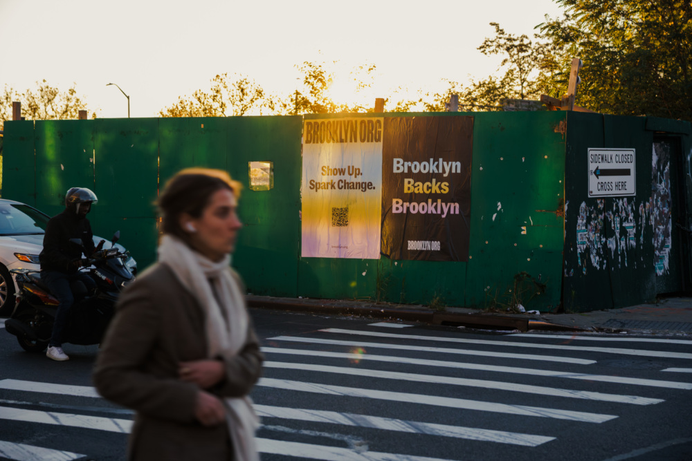 A person crosses a street in front of a green construction barrier with posters reading "Show Up. Spark Change." and "Brooklyn Backs Brooklyn" in the background.