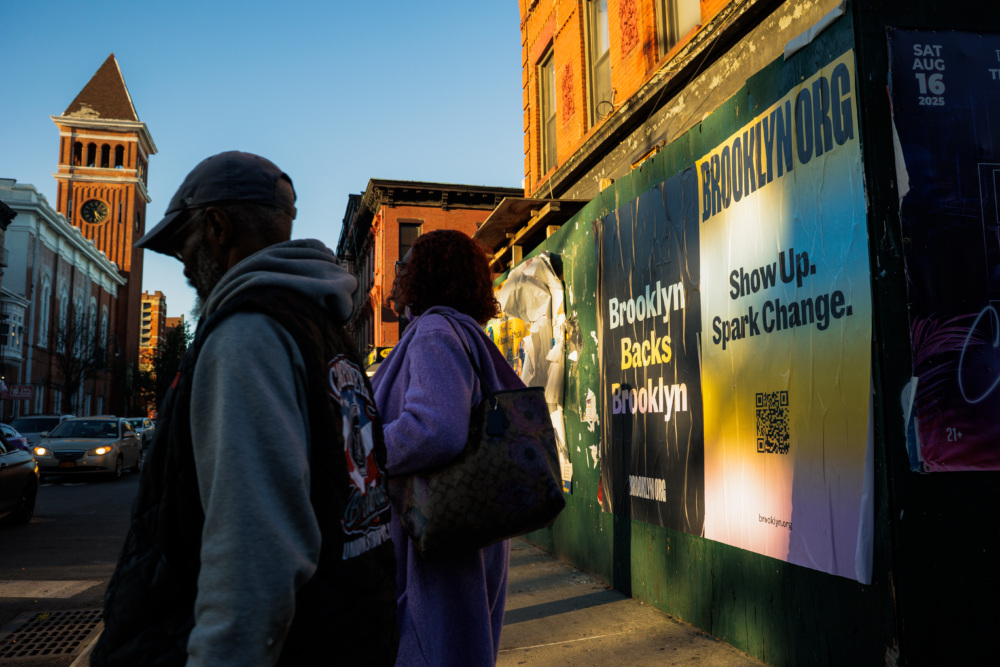 Two people walk past posters on a Brooklyn street at sunset; one poster reads "Brooklyn Backs Brooklyn" and another says "Show Up. Spark Change.