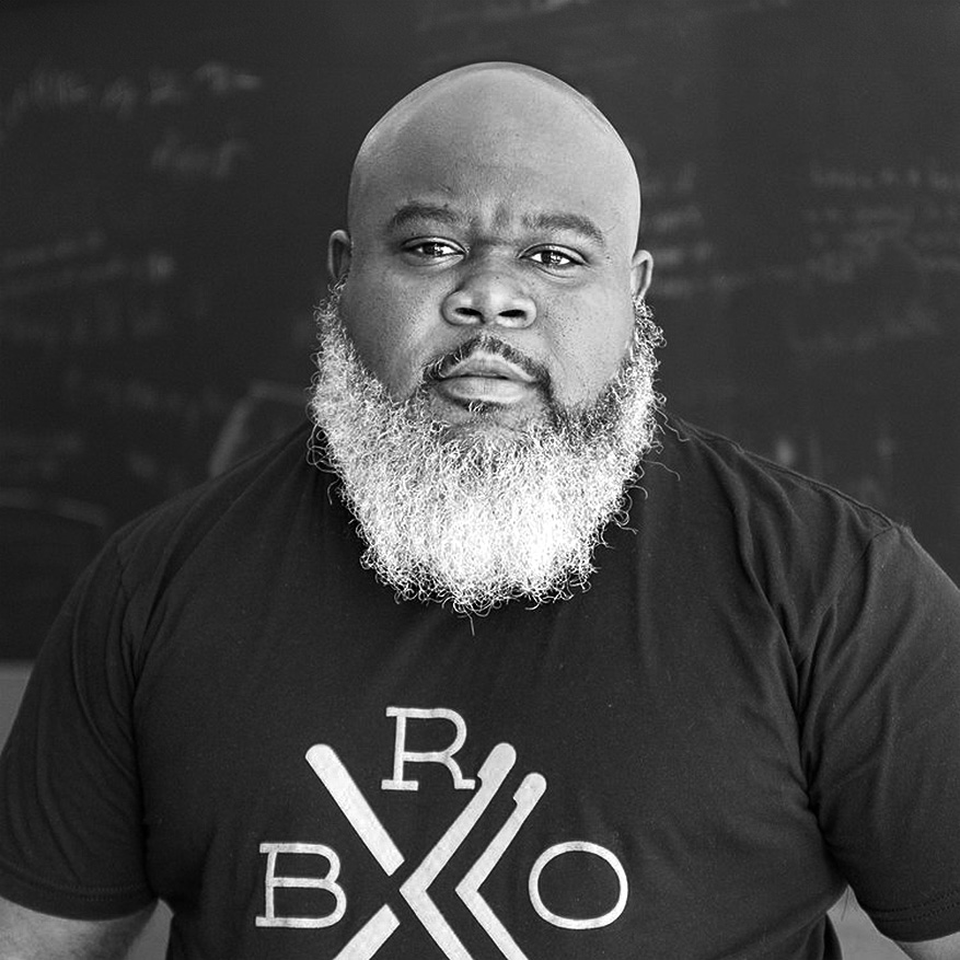 A man with a shaved head and a full, graying beard stands facing the camera, wearing a black t-shirt with white graphic text. A chalkboard with writing is in the background.