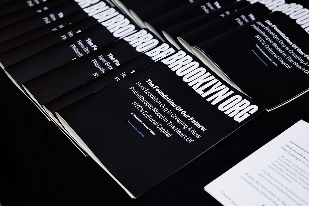 Several black booklets with "BROOKLYN.ORG" on the cover are arranged in a row on a table, alongside a stack of white papers.