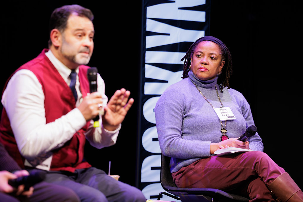 Two people sit on stage during a panel discussion; one speaks into a microphone while the other listens, holding a microphone and notepad.