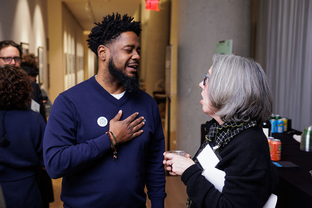 A man and a woman stand indoors having a conversation during an event. The man gestures with his hand on his chest, and beverages are visible on a table in the background.
