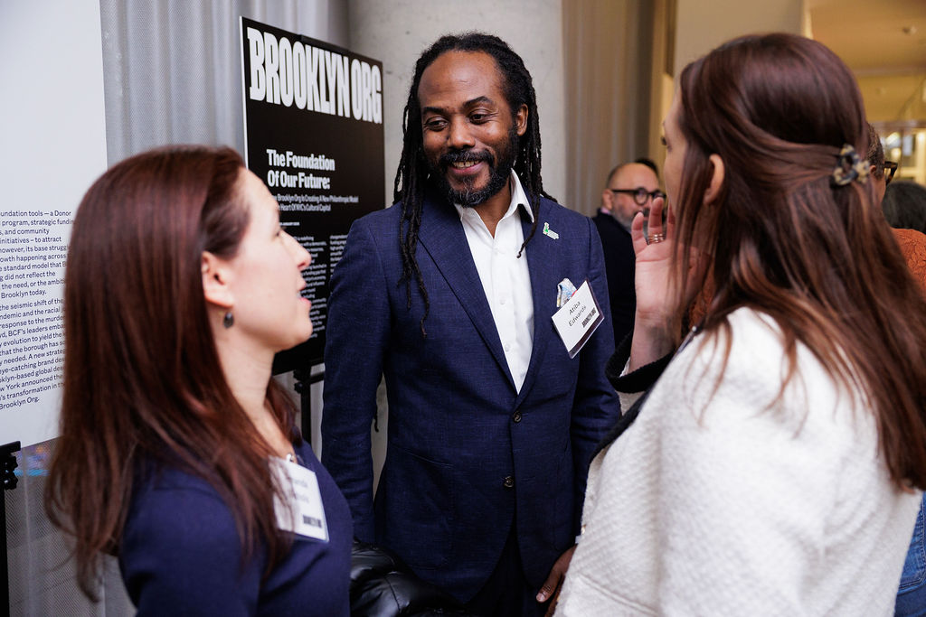 Three people stand and converse at an indoor event. They wear name tags and are dressed in business attire. Informational posters are visible in the background.