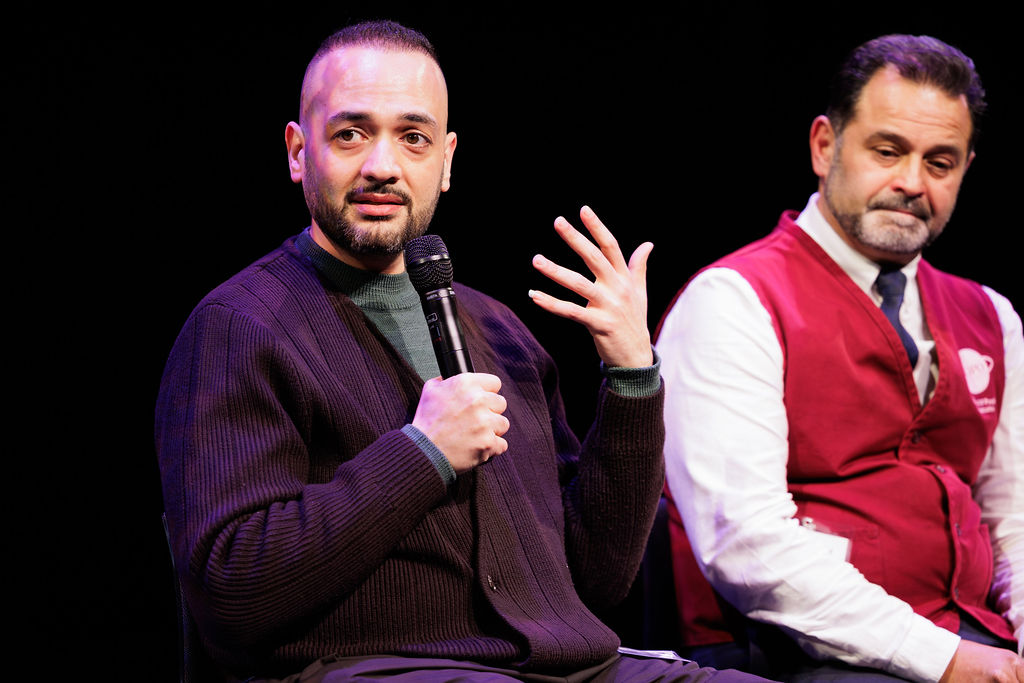 A man speaks into a microphone while gesturing with his hand; another man in a red vest sits beside him, listening attentively against a black background.