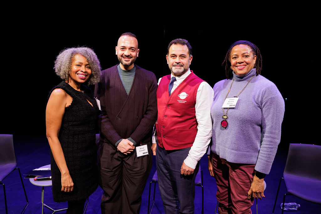 Four people stand side by side and smile at the camera on a stage with empty chairs behind them. They each wear conference name badges.