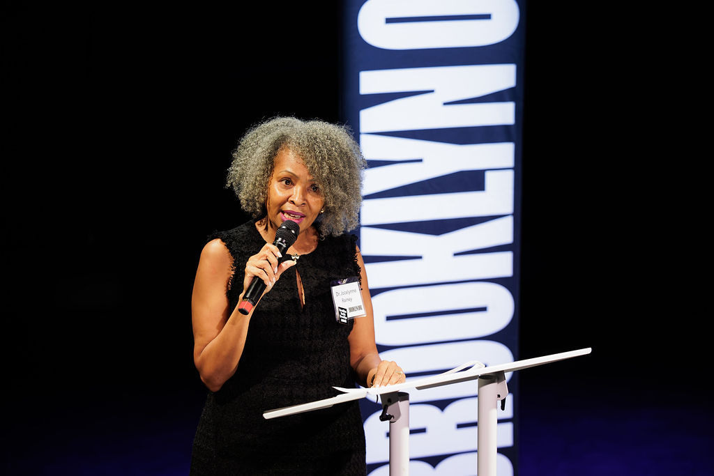 A woman with curly gray hair speaks into a microphone at a podium, with a large vertical "BROOKLYN" sign in the background.