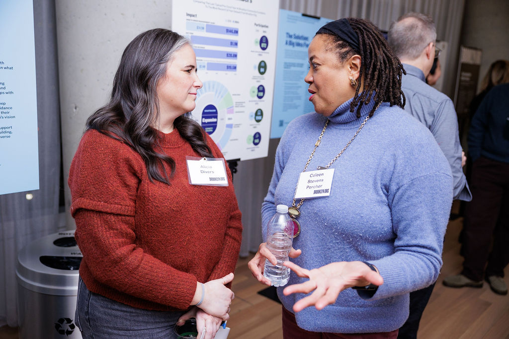 Two women wearing name tags are having a conversation indoors at an event, with informational posters visible in the background.