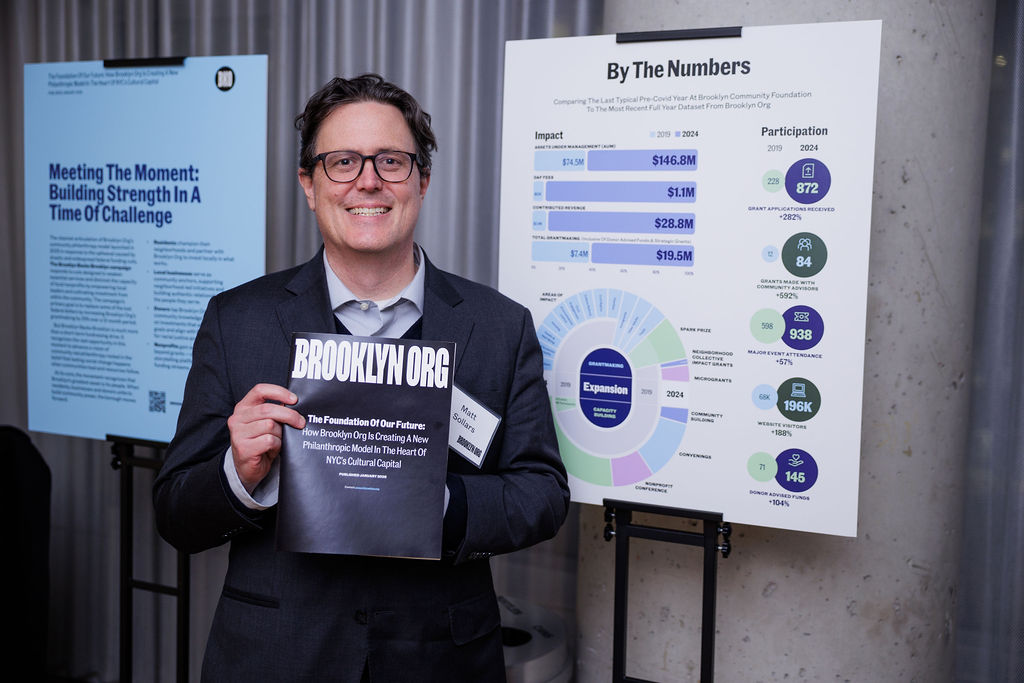 A man wearing glasses and a suit holds a "Brooklyn Org" booklet next to informational display boards with charts and statistics.