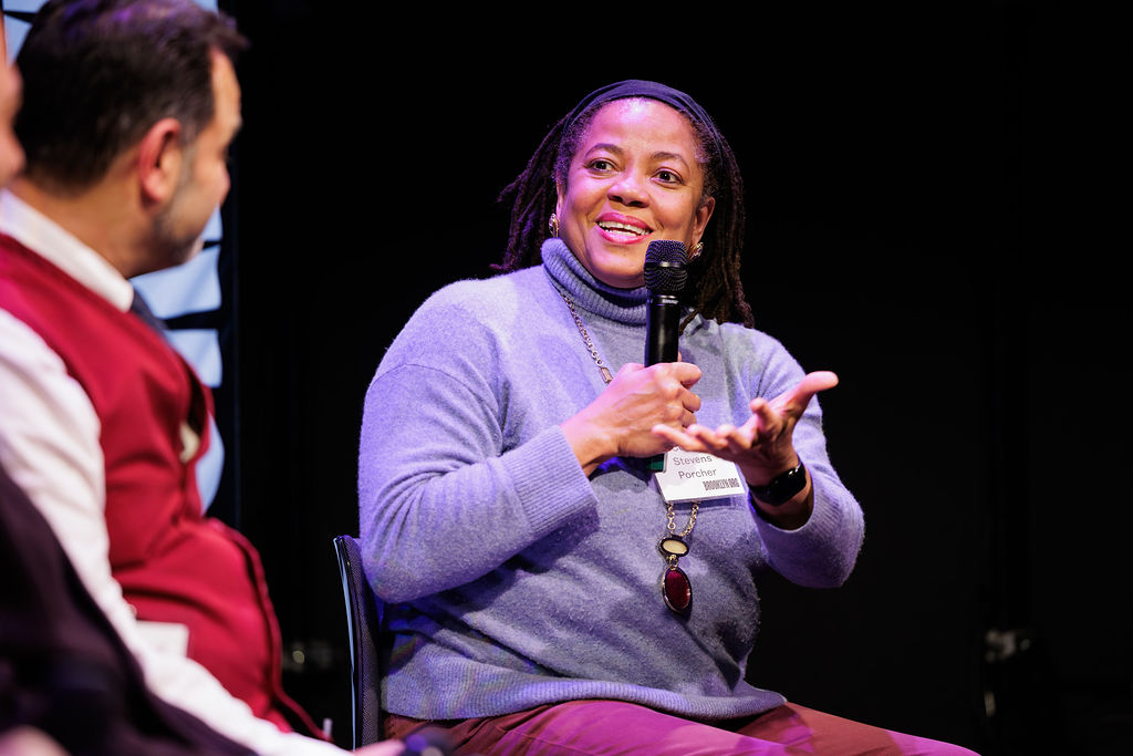 A woman holding a microphone speaks while gesturing with her hand during a panel discussion. Another person, wearing a red sweater vest, is partially visible beside her.