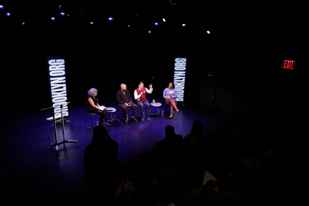 Four people sit on stage under spotlights for a panel discussion, with two vertical screens displaying "BROOKLYN.ORG" in the background.