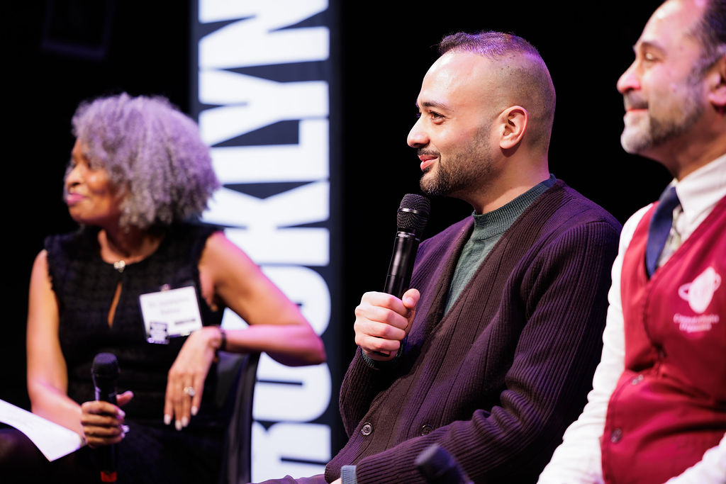 Three people sit on stage holding microphones, engaged in a panel discussion, with a vertical "BROOKLYN" sign in the background.