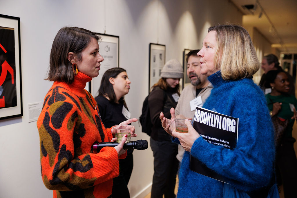 Two women converse at an art gallery event; one holds a microphone, the other holds a drink and a folder labeled "BROOKLYN.ORG." Other attendees are visible in the background.